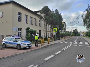 Na zdjęciu policjant i radiowóz w rejonie przejścia dla pieszych przy szkole.
