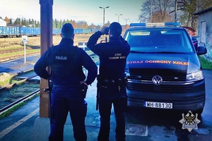 Na zdjęciu policjant oraz funkcjonariusz Straży Ochrony Kolei na dworcu przy radiowozie.