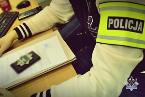 Na zdjęciu policjant po cywilnemu z opaską na ramieniu z  napisem policjant przed nim akta sprawy i odznaka policyjna
