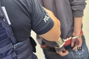 Na zdjęciu policjant pilnuje zatrzymanego mężczyzny w kajdankach.