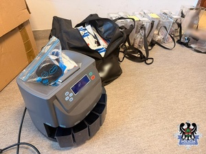 na zdjęciu maszyna do liczenia pieniędzy , torba z zabezpieczonymi pieniądzmi  zapakowanymi w koperty i zabezpieczone puszki z pieniędzmi