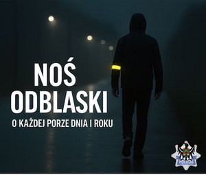 Zdjęcie przedstawia sylwetkę mężczyzny idącego ulicą nocną porą z widocznym elementem odblaskowym na ramieniu i napis Noś odblaski badż widoczny