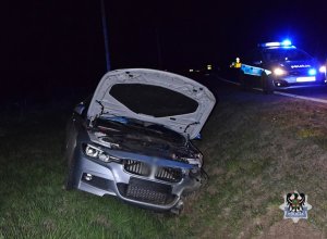 Na zdjęciu uszkodzenia samochodu, którym kierował nietrzeźwy kierujący, w tle radiowóz - ujęcie z przodu.